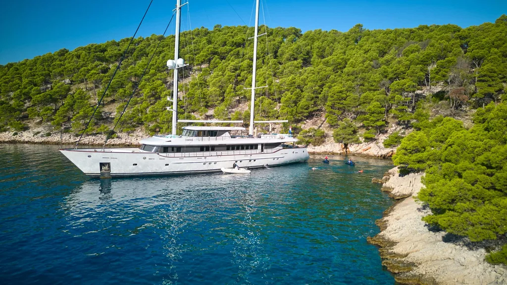 OMNIA anchored near a pine-covered coastline in clear blue water.