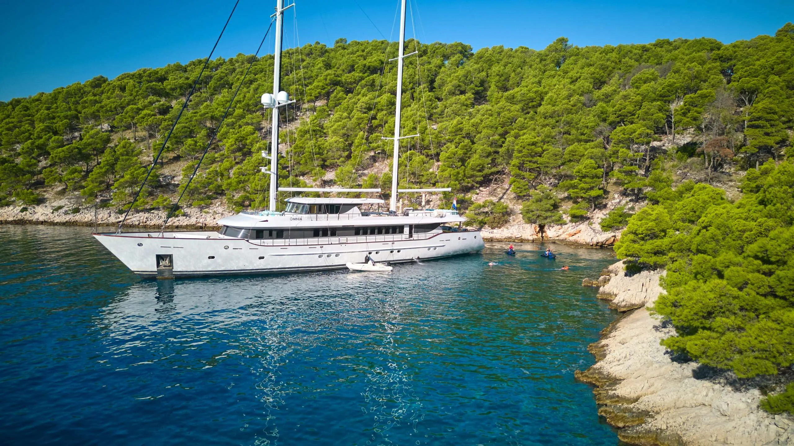 OMNIA anchored near a pine-covered coastline in clear blue water.